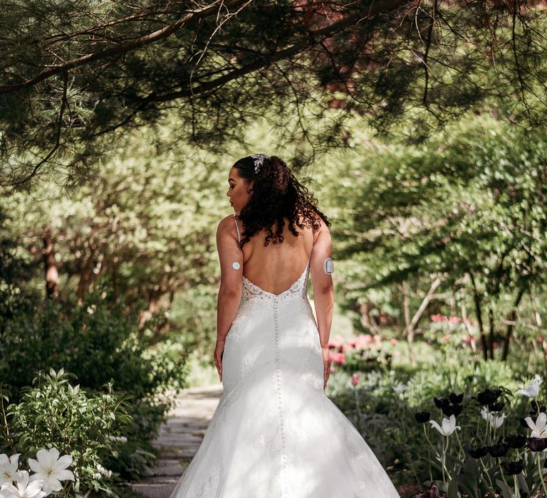 Bride stands within gardens and wears wedding gown with V-shaped back and straps