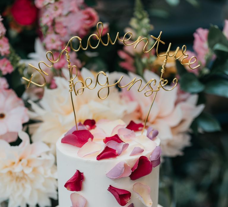 Two-tier wedding cake complete with white frosting, petal decoration in different tones of pink and golden cake topper quote