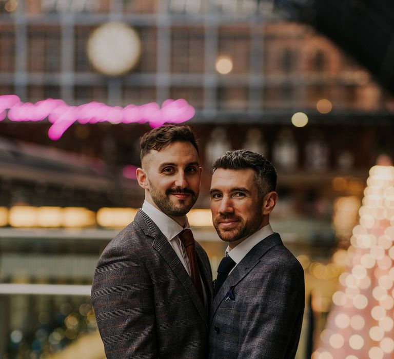 Grooms look towards the camera as they hold one another one their wedding day in front of pink neon sign