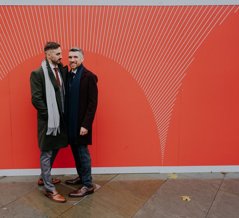 Grooms stand in front of bright red wall outdoors as they lean into one another on their wedding day in London