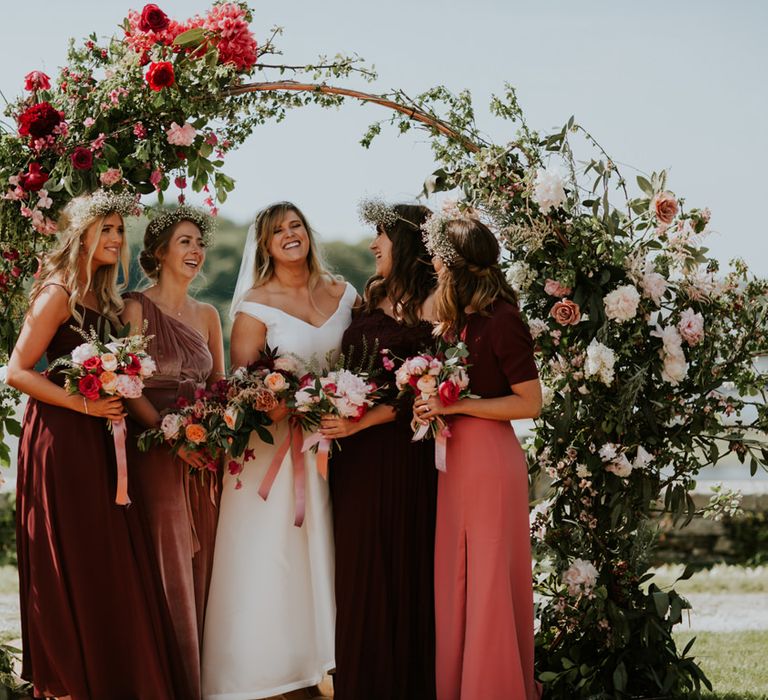 Bride and bridesmaids portrait by a floral arch in different style and fabric burgundy dresses for outdoor summer wedding