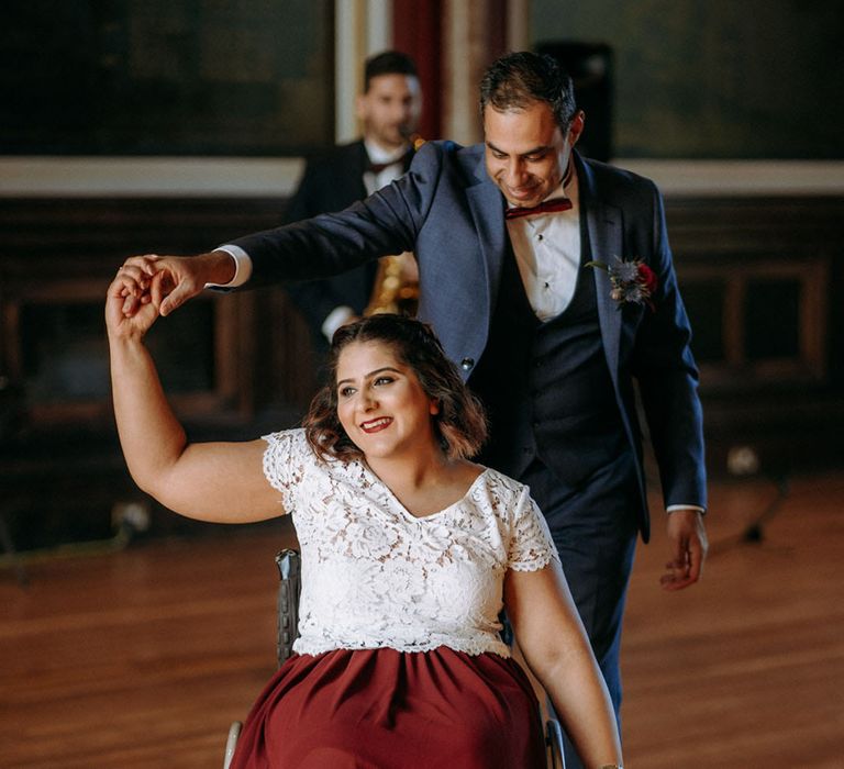 Bride and groom first dance at Dulwich College with bride in a wheelchair wearing a lace top and burgundy top as her groom twirls her in a blue suit and bow tie 