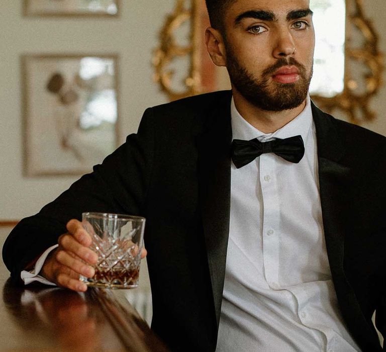 Groom in a tuxedo drinking whisky at the bar at Bourton Hall wedding venue 