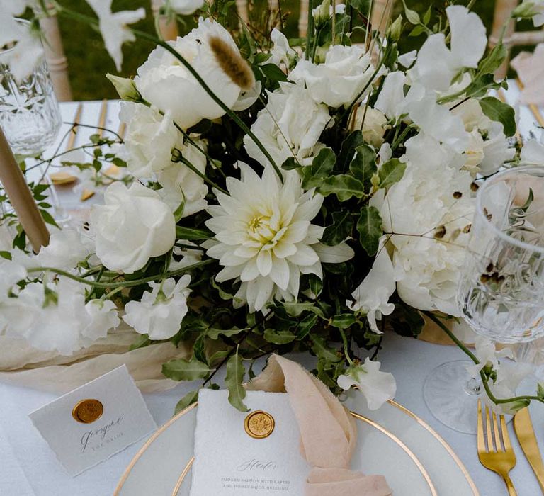 Place setting with gold rimmed tabled ware, gold cutlery and white and green floral centrepiece