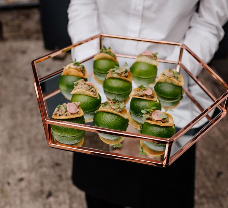 Waitress in long sleeved white shirt holding hexagonal gold rimmed mirrored tray with round green canapés during wedding reception at Loft Studios London