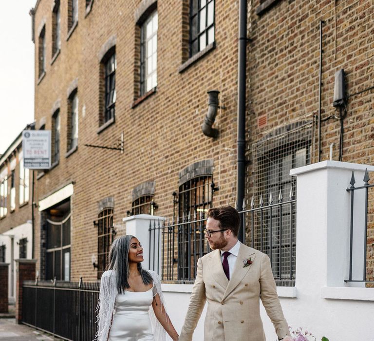 Bride in satin Halfpenny London wedding dress and tasselled bridal cape holds hand with groom in double breasted lien suit and gold trainers holding multicoloured wedding bouquet as they walk along street after wedding ceremony at Loft Studios London