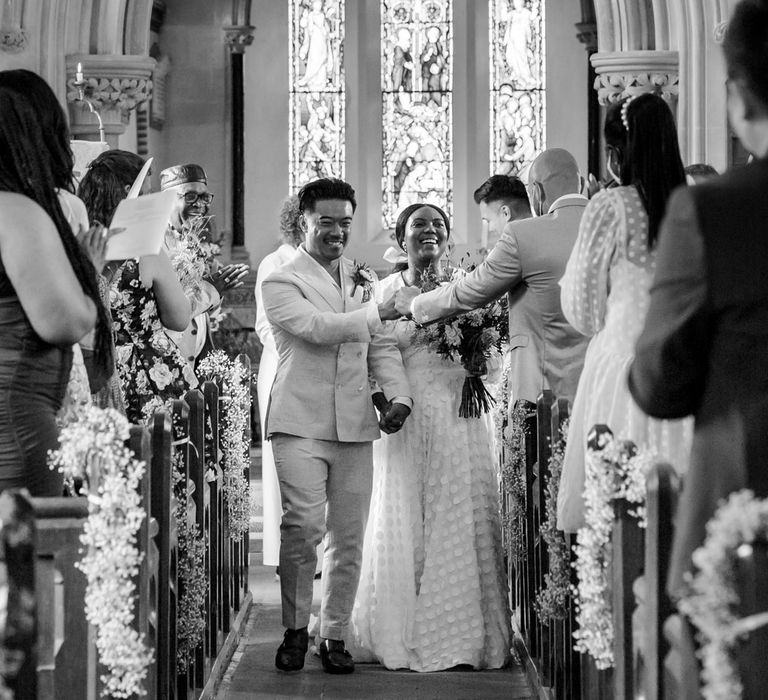 Bride in long mesh sleeve spotted Vagabond wedding dress holding mixed bridal bouquet and groom in double breasted linen suit walk down the aisle after church wedding ceremony at Wasing Park