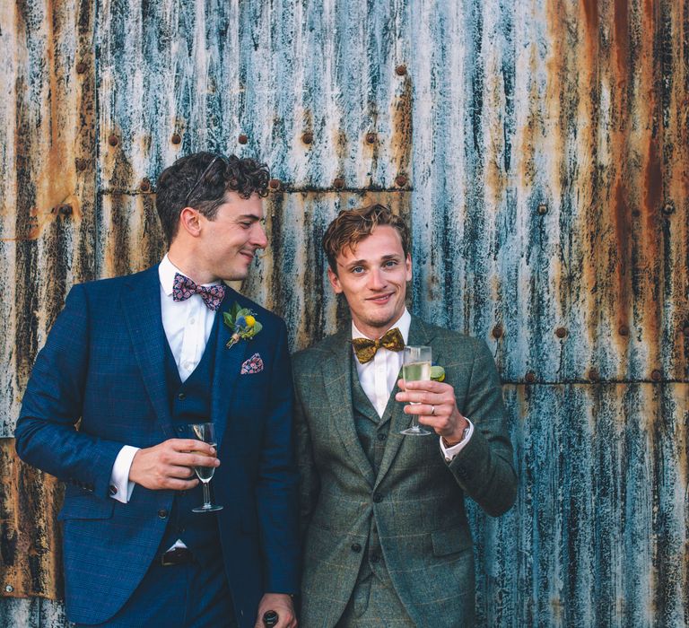 Grooms stand beside industrial style wall as they drink and laugh with one another  | Story + Colour