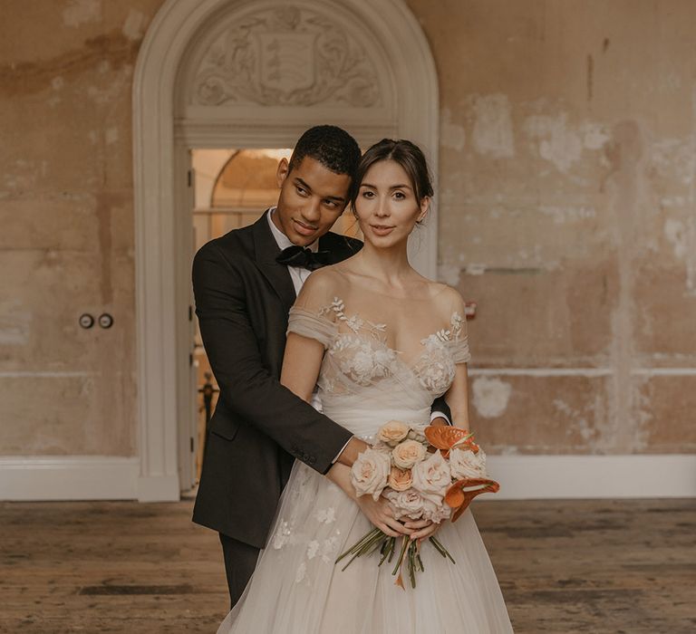 Groom in a black tuxedo embracing his bride in a tulle Bardot wedding dress with illusion neckline and lace detail 