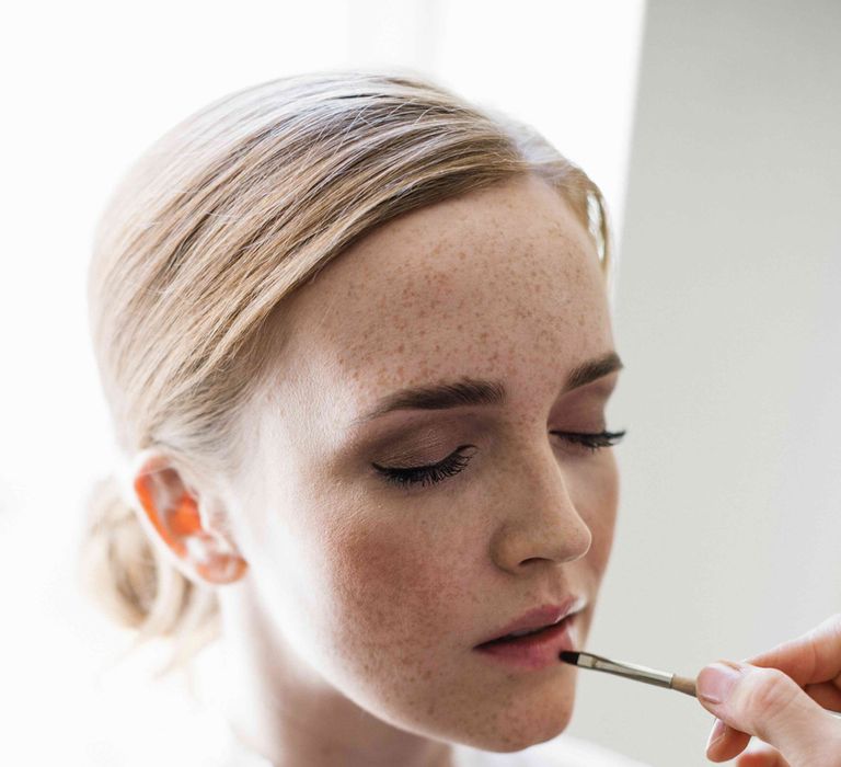 Blonde bride with freckles and swept back hair applying her natural wedding makeup 