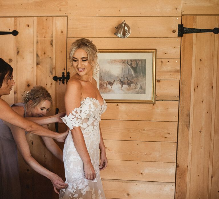 Bride with curled wedding updo has off the shoulder Enzoani wedding dress done up by bridesmaids in purple bridesmaid dresses before tipi wedding at Inkersall Grange Farm