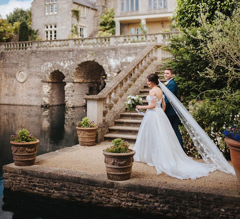 Bride stands besides steps on her wedding day in Suzanne Neville gown and Martina Liana veil