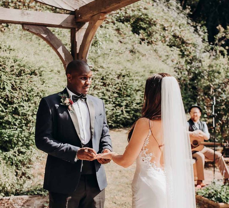 Bride in Scope back with lace detail stands with Groom in dark suit, black bow tie & white shirt in sunny outdoor ceremony 