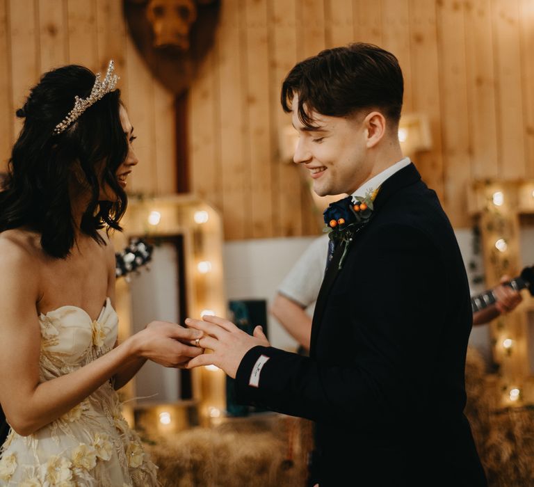 Bride in strapless feathered wedding dress and headpiece puts rings on grooms finger as he wears blue suit and floral buttonhole at rustic wedding in Lancashire