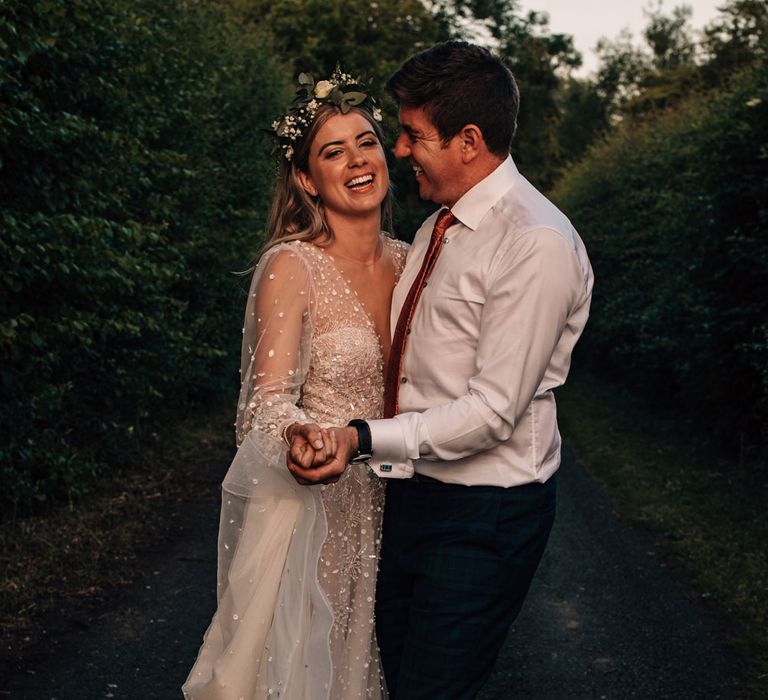 Bride in boho flower crown and long sleeve Julie Vino wedding dress smiles whilst hugging groom in white shirt during golden hour