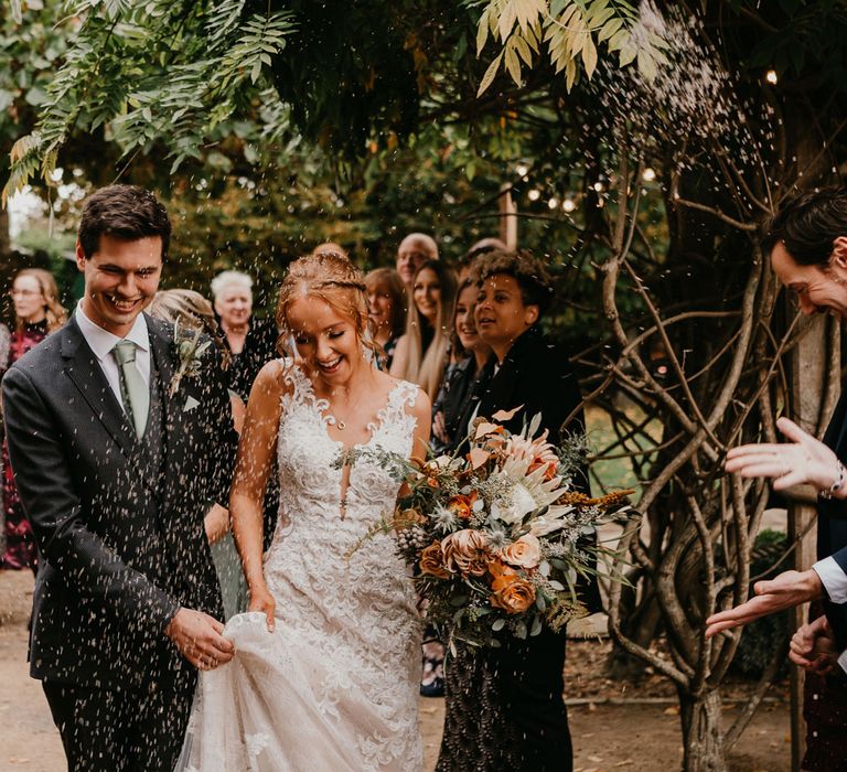 Groom in dark suit and light blue tie holds tulle skirt of brides wedding dress as guests throw multicoloured confetti after rustic wedding ceremony in barn