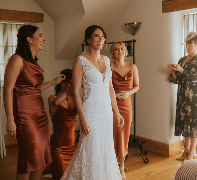 Bride in lace V neck wedding dress with train stands with bridesmaids in orange satin bridesmaid dresses before wedding