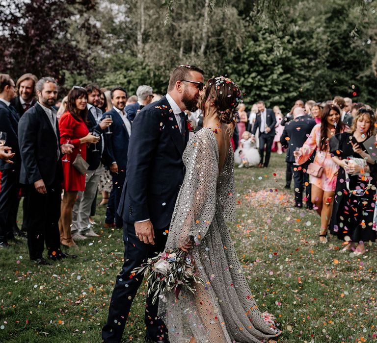 Bride in a silver sequin wedding dress and groom in a navy suit kissing at the end of the confetti line 