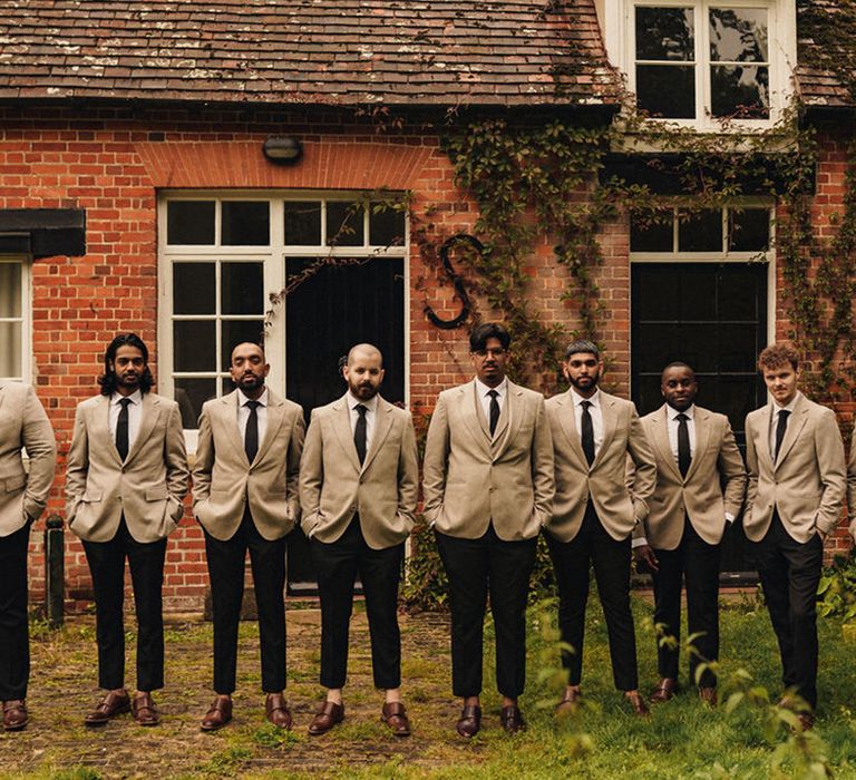 Groom stands with his groomsmen who wear beige suits complete with black ties and trousers and brown brogues 