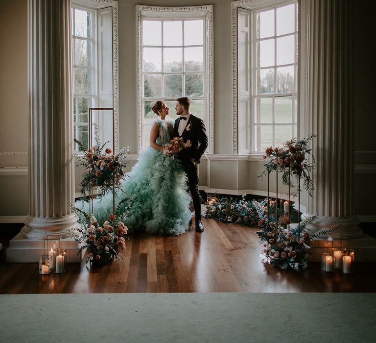 Bride in a Millia London teal wedding dress and groom in a tuxedo standing in front of the windows at Newburgh Priory with metal stands decorated in flowers around them 