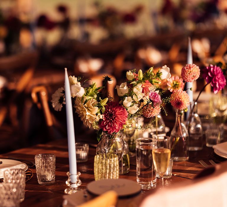 Bright florals in glass vases on wooden table in marquee for garden wedding reception with tambourine place names, light blue candles and textured glassware