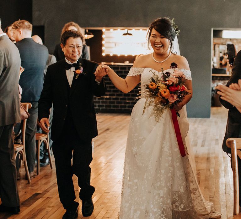 Bride in lace off the shoulder wedding dress and pearl necklace holding pink and red bridal bouquet walks down the aisle holding hands with father of the bride in black tie and colourful floral buttonhole