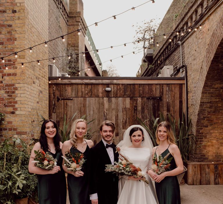 Bride in long sleeve wedding dress and veil stands with groom in black tie and bridesmaids in dark green bridesmaid dresses