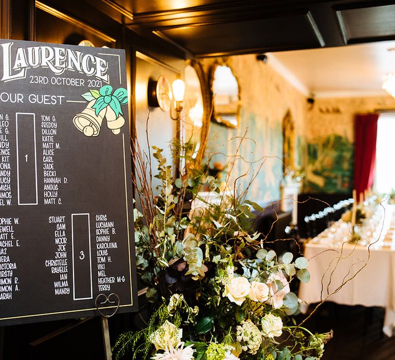 Black board wedding sign of seating chart plan with large flower display of roses and green foliage