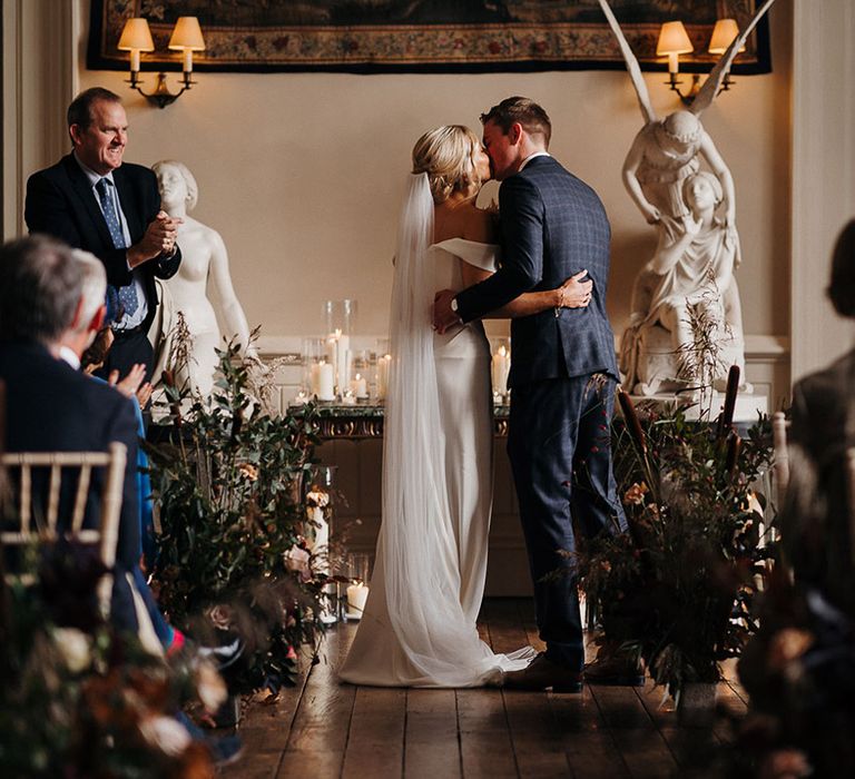 Groom in blue checkered suit and bride in off the shoulder wedding dress share their first kiss as a married couple