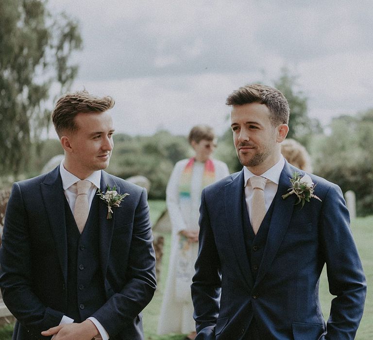 Groom and groomsman in matching three-piece navy blue suit and beige ties with white flower buttonholes 