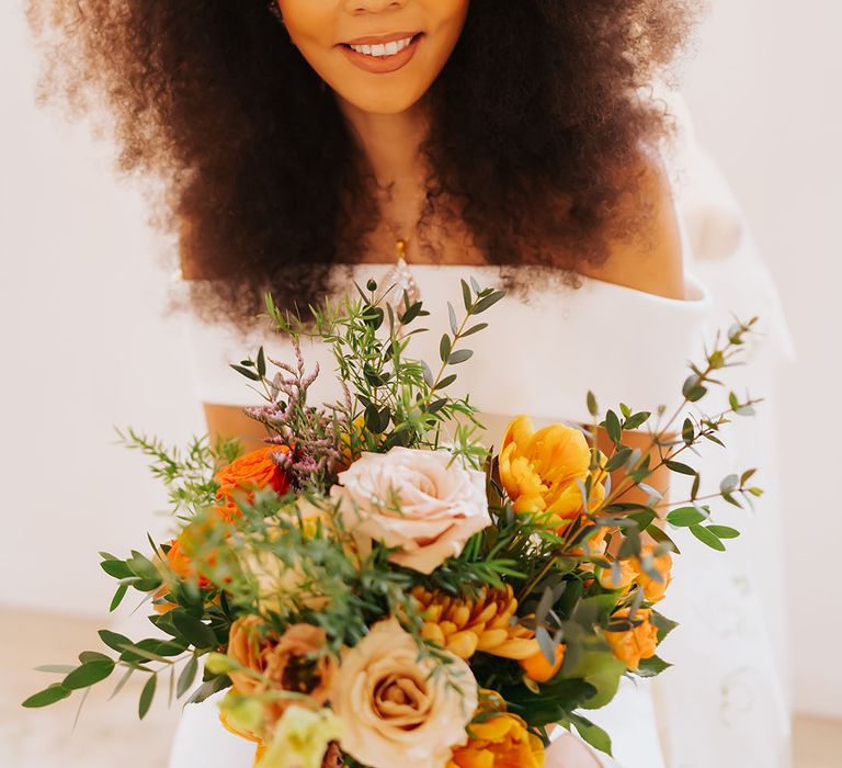 Black bride with afro hair wearing a hair accessory and holding an orange, yellow and white golden wedding bouquet 