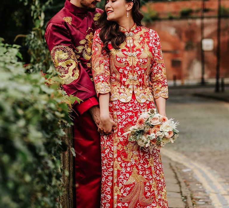 Bride and groom in red and gold traditional Chinese wedding attire with bride holding autumnal wedding bouquet