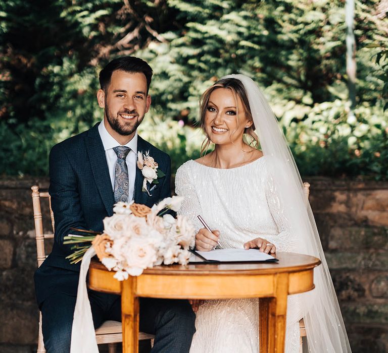 Groom in dark blue suit with paisley tie signs the register with bride in beaded wedding dress