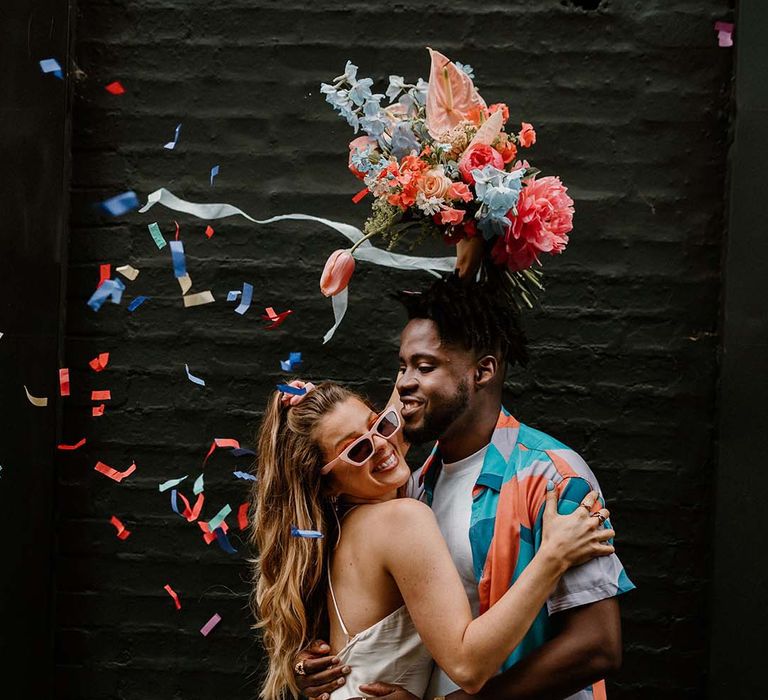 Colourful confetti moment at city elopement with bride in a short wedding dress holding a pink and blue bouquet in the air and groom in a patterned shirt and white tee