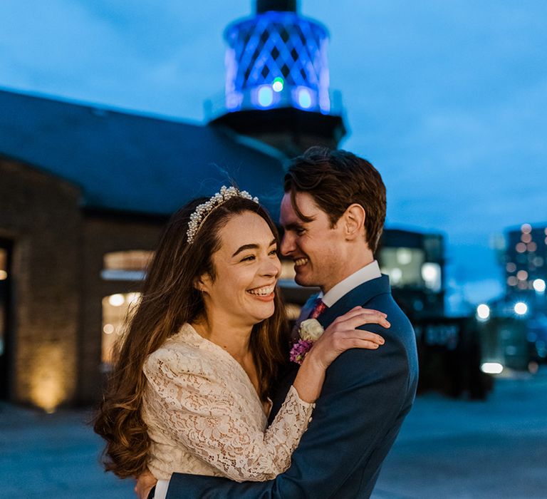 Bride in lace dress and pearl cluster headband hugs groom in blue suit and pink and blue patterned tie at their city party wedding