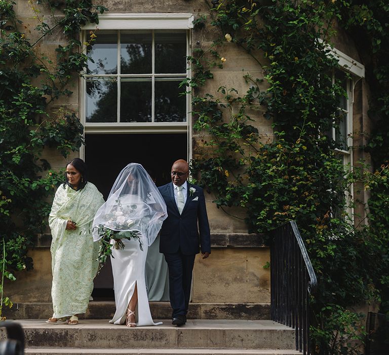 Father and mother of the bride walk their daughter down the aisle with veil and off the shoulder wedding dress 