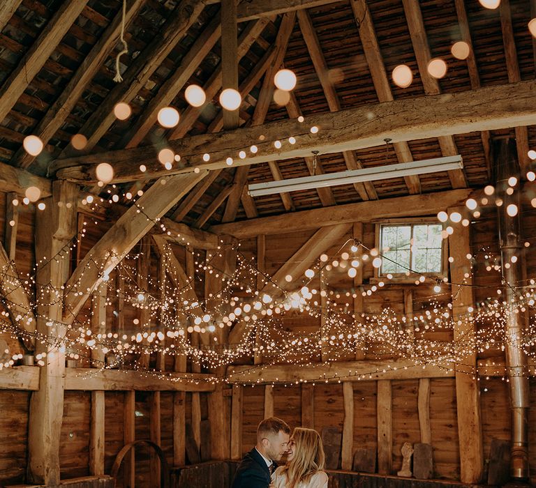 Bride in beaded wedding dress and groom in blue suit hold each other under twinkling fairy lights at The Secret Barn