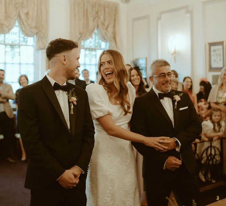 Bride smiles widely at groom as her father walks her down the aisle for their registry office wedding