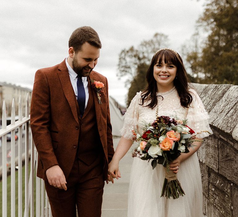 Bride and groom hold hands as they pose together around the city of York with bride wearing gold headband and groom in brown suit with a blue tie