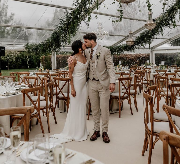 Bride in a satin wedding dress and groom in a beige suit kissing in the middle of their clear glass marquee wedding reception with wooden furniture, greenery decor and handing chandelier 