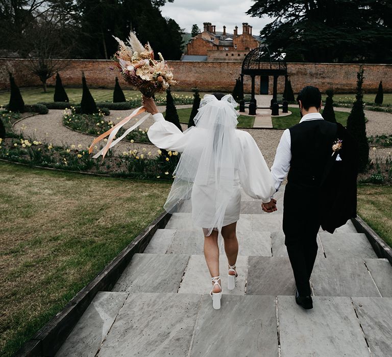 Bride in a short wedding dress with long sleeves wearing platform shoes, and a bow veil holding hands with her groom in a tuxedo 