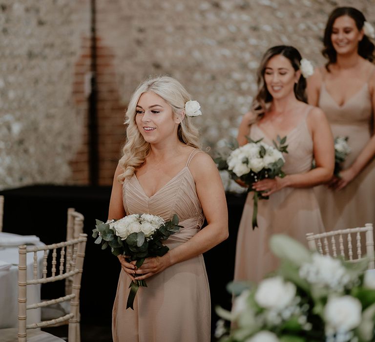 Bridesmaids in champagne pink dresses walk down the aisle together carrying white rose bouquets with matching flowers in their hair 