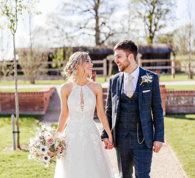 Bride in floral tulle halter neck wedding dress walking holding hands with groom in blue checker suit and pale tie with gold tie clip 