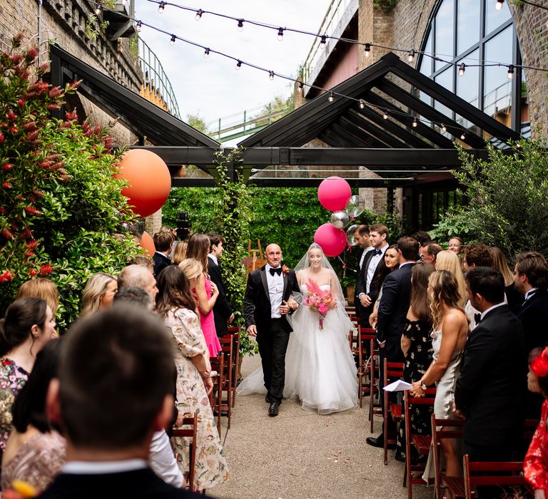 Bride walks down the aisle lined with orange and pink balloons as she holds colourful dried floral bouquet with pink pampas grass