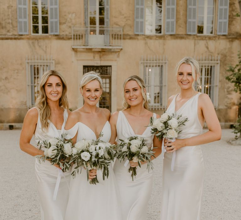 Bride wearing Suzanne Neville wedding dress stands with her bridesmaids who wear silk white bridesmaids dresses with cowl necks and carry white rose bouquets