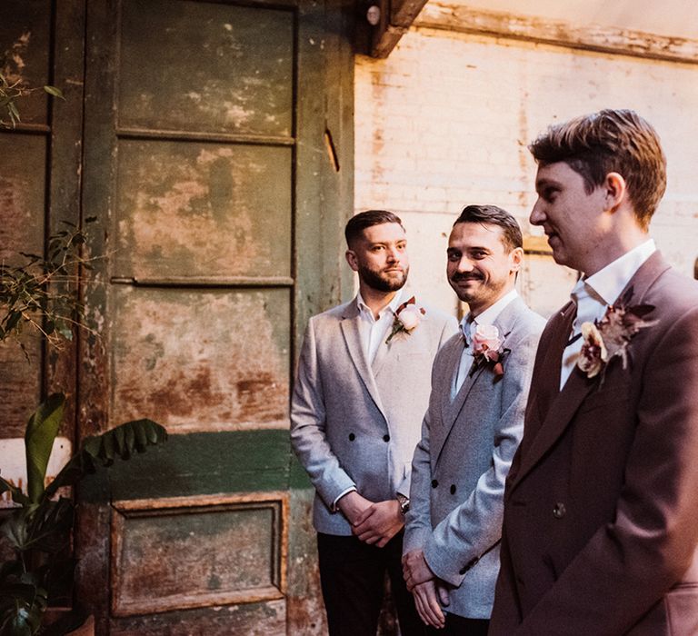 Best men in grey suits smile at the groom who wears a brown suit and large flower buttonhole 