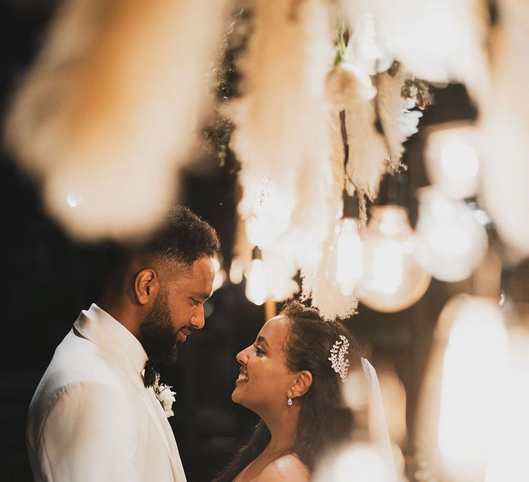 Bride & groom look lovingly at one another surrounded by pampas grass bouquets and festoon lighting 