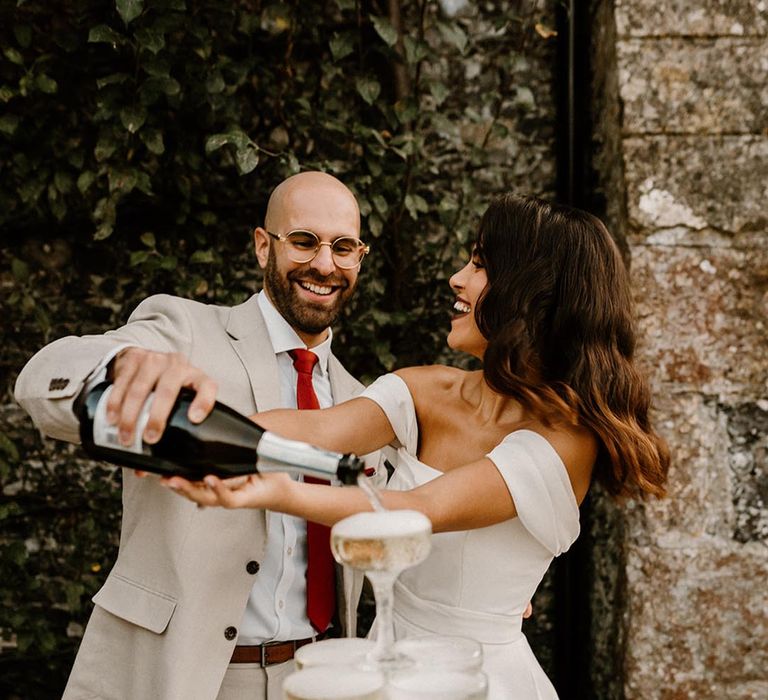 Bride and groom smile brightly as they pour champagne onto their champagne tower 