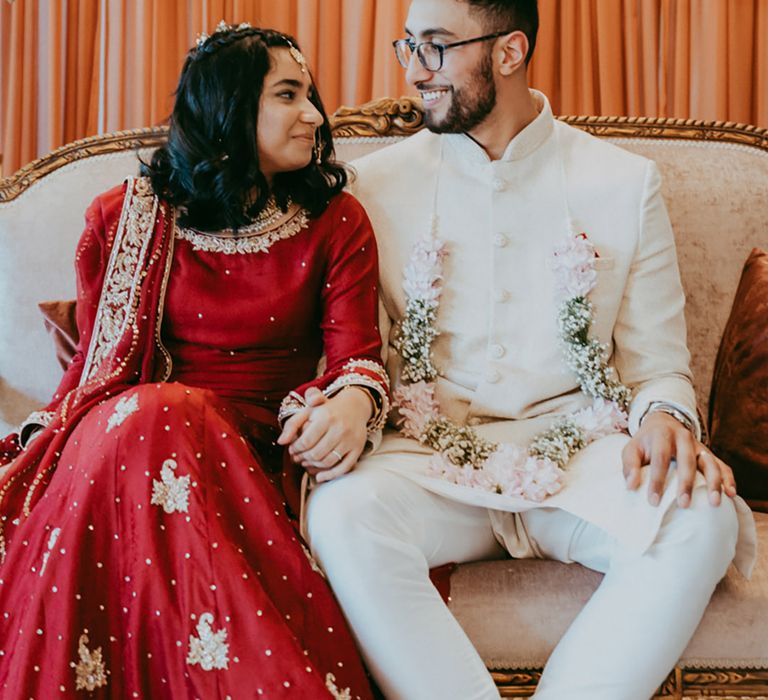 Bride in red and gold embellished bridal two-piece looks lovingly at her groom who wears pastel lei 