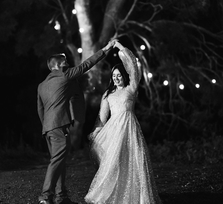 Bride & groom dance outdoors surrounded by festoon lighting in black & white image